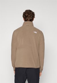Fleece jacket in tan with a high collar, long sleeves, and a small white logo at the upper back. Soft texture with a relaxed fit.