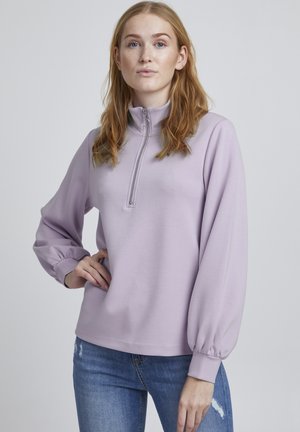 Lys lilla pullover med halv-zip krave, flagrende lange ærmer og ribbede manchetter. Glat tekstur og afslappet pasform, parret med slidte jeans.