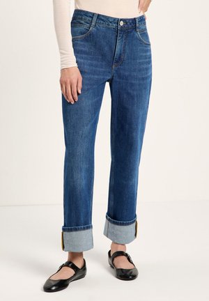 Person mit hochgekrempelten blauen Jeans, einem hellen langärmligen Oberteil und schwarzen Mary Jane Schuhen, stehend auf einem weißen Boden.