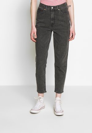 Femme portant un jean en denim noir taille haute, des baskets blanches montantes et un t-shirt à rayures rouges et blanches, debout devant un fond uni.