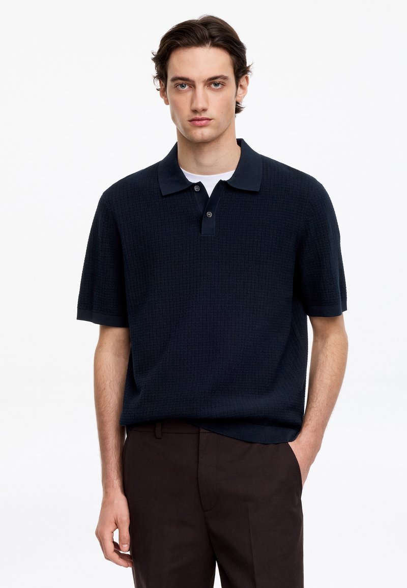 Jeune homme aux cheveux bruns courts portant un polo noir texturé à manches courtes et un pantalon marron foncé, debout avec une main dans la poche.
