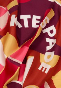 Μεταξωτό μαντήλι με γεωμετρικό σχέδιο σε αποχρώσεις κόκκινου, ροζ, πορτοκαλί και κίτρινου, με έντονο λευκό κείμενο που εμφανίζει το όνομα της μάρκας.