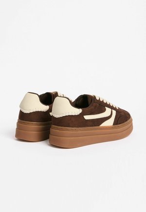 Bruine suède sneakers met crèmekleurige accenten, verhoogde rubberen zool en gestikte details op de hiel. Veters en zijkantlogo in een contrasterende kleur.