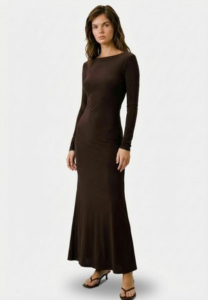 Nő áll, visel egy testhezálló, hosszú ujjú, földig érő sötétbarna ruhát és fekete, nyitott orrú, magas sarkú szandált egyszínű háttér előtt.