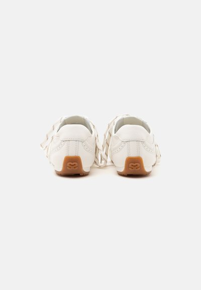 Paire de chaussures en cuir blanc avec semelles en caoutchouc beige vue de dos, montrant des détails de couture perforée et un logo décoratif.