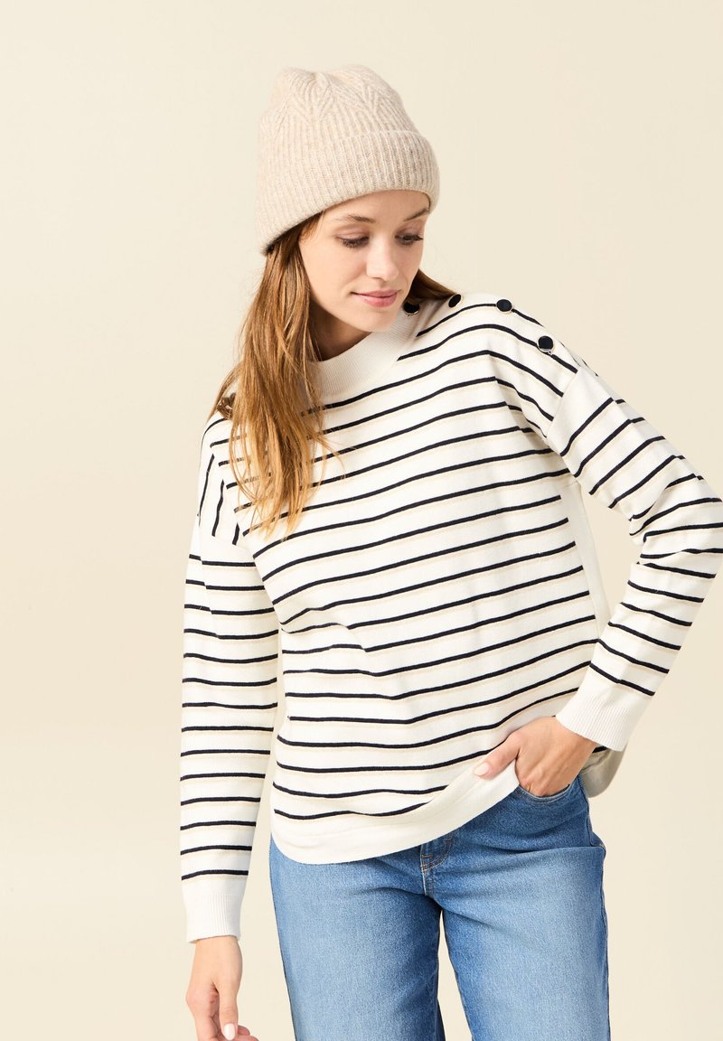 Pull en tricot rayé crème avec des lignes horizontales navy, doté d'un col rond et d'accents de boutons aux épaules. Porté avec un jean bleu clair.