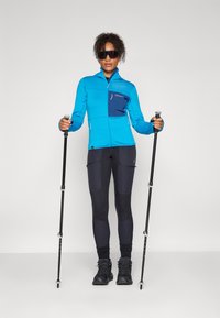 Blå zip-up jakke med teksturert stoff, marineblå lomme, sorte leggings og sorte turstaver. Solbriller fullfører utendørslooken.