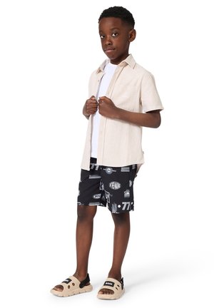 Garçon debout avec assurance, portant une chemise beige à manches courtes sur un T-shirt blanc, un short noir imprimé et des sandales slides beiges avec des sangles noires.