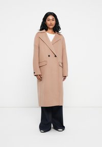 Cappotto in lana beige doppiopetto con colletto a revers, vestibilità oversize e bottoni neri, abbinato a jeans neri a gamba larga.