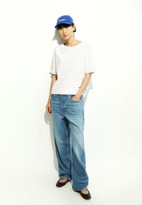 Striped white and light blue t-shirt, relaxed fit, paired with wide-leg light blue jeans. Black flat shoes and a blue cap complete the look.