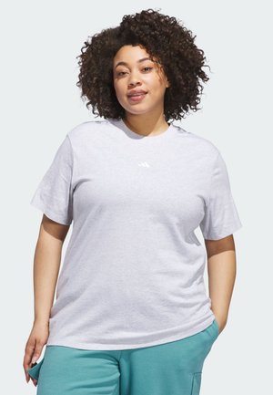 Ung kvinde med krøllet hår iført en lysegrå Adidas t-shirt og turkis bukser stående foran en hvid baggrund.