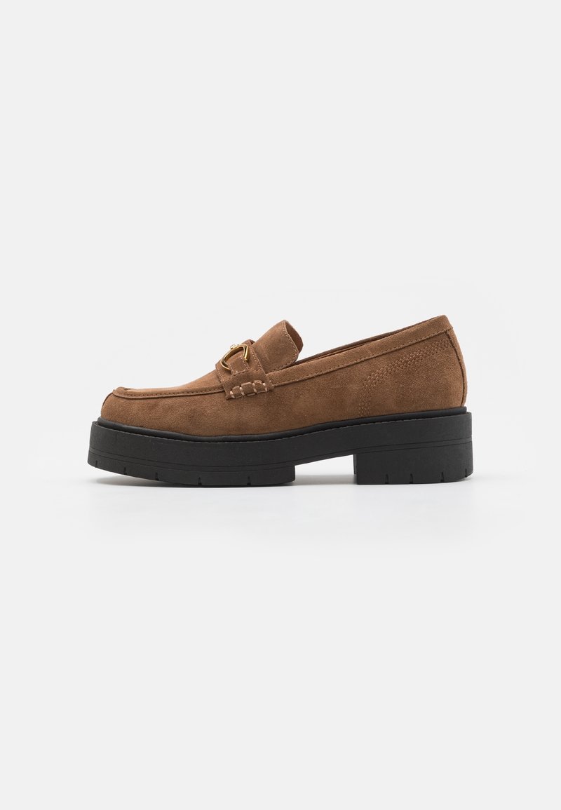 Mocassin en daim marron avec semelle noire épaisse et détail boucle dorée sur le dessus, présenté en vue latérale sur fond blanc.