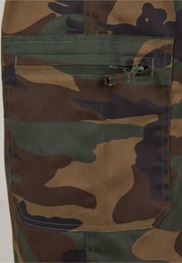 A kamuflázs cargo nadrágok zöld, barna és fekete minták keverékéből készültek. Cipzáras zseb és erősített varrás jellemzi.
