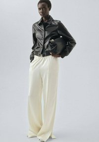 Kobieta w czarnej skórzanej kurtce, w szerokich kremowych spodniach, trzymająca dużą czarną kopertówkę na tle jasnego, jednolitego tła.