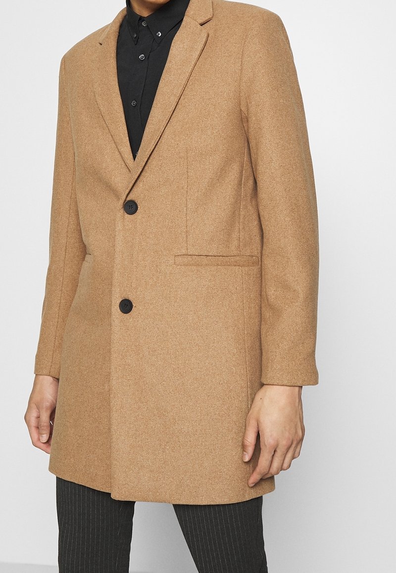 Cappotto in misto lana marrone con vestibilità su misura, design a un bottone, rever a vista e chiusure con bottoni neri; abbinato a una camicia nera.