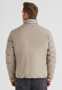 Beige gepolsterte Jacke mit einem hohen Kragen, elastischen Bündchen und einer subtilen Textur. Verfügt über Branding auf der Rückseite und wird mit dunklen Jeans kombiniert.