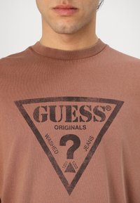Brūns, īsu piedurkņu T-krekls, izgatavots no kokvilnas. Tam ir trīsstūra melns grafiskais attēls ar uzrakstu "GUESS ORIGINALS" un jautājuma zīmes dizains.
