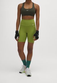 Sujetador deportivo verde caqui, pantalones cortos de cintura alta en verde lima, guantes de entrenamiento negros y calcetines verdes combinados con zapatillas deportivas gris claro.