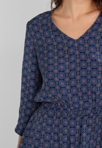 Robe bleue à motif avec un décolleté en V, ornée de motifs circulaires complexes en rose, blanc et bleu, taille froncée et manches longues.