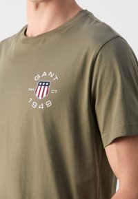 Olivgrön bomullst-shirt med rund halsringning, med en cirkulär logotyp i rött, vitt och blått och året "1949" under.