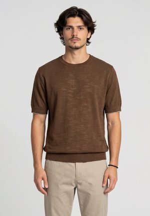 Giovane uomo con capelli castani di media lunghezza, che indossa un maglione marrone a maniche corte e pantaloni beige chiari, in piedi davanti a uno sfondo semplice.