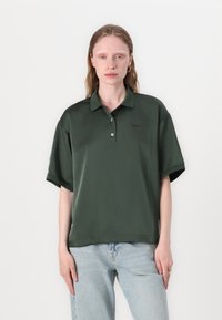 Mørkegrønn polo skjorte med korte ermer, krage og en subtil logo p�å brystet, kombinert med lyseblå jeans. Glatt tekstur.