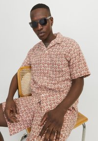 Kurzarmhemd und Short-Set in Hellbraun mit geometrischen Mustern, aus leichtem Stoff gefertigt. Modell trägt Sonnenbrille und sitzt auf einem Stuhl.