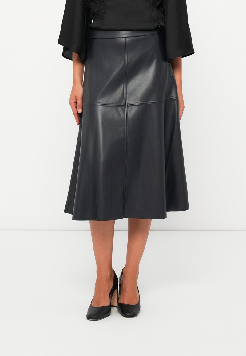 Jupe midi en cuir noir avec détails des coutures, portée avec des talons hauts noirs et un top noir.