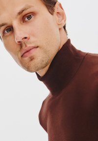 Brązowy sweter z golfem, wykonany z gładkiego materiału, z żeberkowym kołnierzem. Krój jest dopasowany, podkreślający szyję i ramiona.
