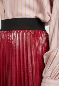 Roter plissierter Rock mit glänzender Oberfläche, ausgestattet mit einem schwarzen elastischen Bund und einem goldenen Herzanhänger mit der Aufschrift "Twinset". Teilweise sichtbare pinkgestreifte Bluse.