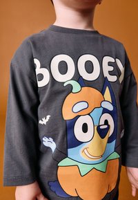 Graues Langarm-T-Shirt mit einer Zeichentrickfigur, die einen Kürbishut trägt und große Augen hat, zusammen mit dem Wort "BOOEY." Fledermaus-Grafik inklusive.