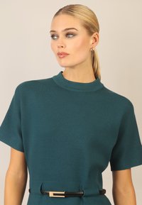 Vestido corto de color teal con manga corta y cuello alto, que presenta una cintura ajustada y un cinturón negro con una hebilla rectangular dorada. Textura suave.