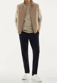 Béžový vest přes světle béžový svetr, v kombinaci s tmavými kalhotami a bílými teniskami. Hladká textura s volným střihem a bez viditelných vzorů.