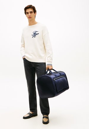 Junger Mann in weißem Pullover und schwarzer Jeans, der eine dunkelblaue Reisetasche vor einem schlichten weißen Hintergrund hält.