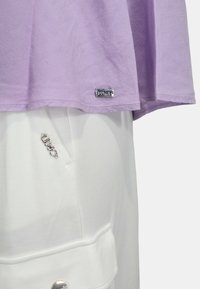 Camicia a maniche corte lavanda con orlo cucito e etichetta in metallo; abbinata a pantaloni bianchi con un accento di gioielli e dettaglio della tasca.