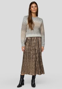 Hellgrauer und blauer Strickpullover mit einem gerippten Halsausschnitt. Metallischer, plissierter bronzefarbener Rock. Modell trägt schwarze Stiefeletten und steht vor einem grauen Hintergrund.