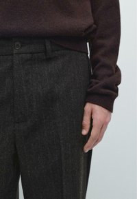 Pantaloni in lana scura con una sottile riga, abbinati a un maglione bordeaux. I pantaloni presentano una chiusura a bottone e due tasche laterali.