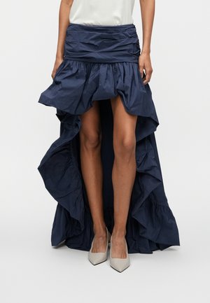 Femme portant une jupe asymétrique bleu marine à volants et des talons blancs pointus, debout devant un fond uni.