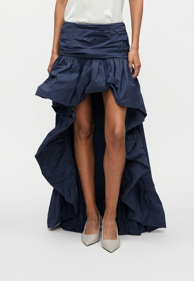 Femme portant une jupe asymétrique bleu marine à volants et des talons blancs pointus, debout devant un fond uni.