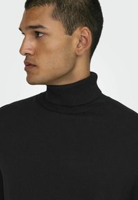 Zwarte coltrui gemaakt van een zachte gebreide stof. Heeft een hoge kraag en een geribbelde textuur rond de nek. Eenvoudig, strakke ontwerp.