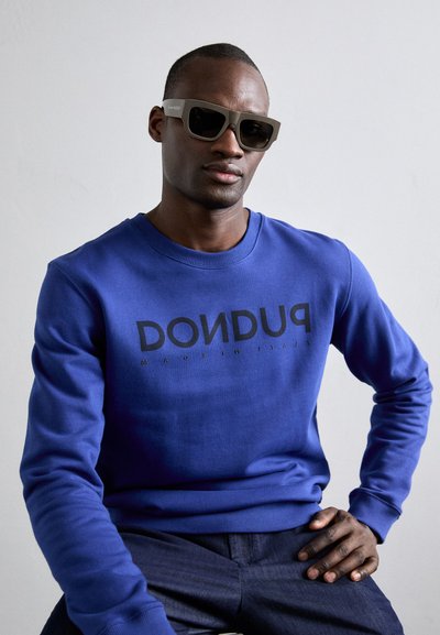 Blue sweatshirt with a black printed logo, crew neckline, and ribbed cuffs, paired with angular, dark sunglasses.