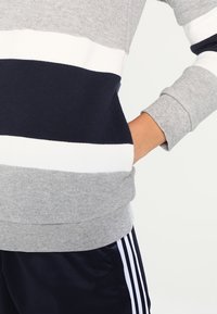 Grå, marinblå och vit randig sweatshirt med ribbad krage och muddar; mjuk textur; lagerdesign; sidofickor synliga.