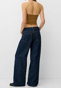 Donna che indossa un top marrone senza spalline e jeans a gamba larga di colore blu scuro, in piedi con le mani nelle tasche, rivolta lontano dalla macchina fotografica.