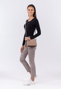 Borsa a tracolla beige in materiale testurizzato, con un logo discreto. Il modello indossa un top nero a maniche lunghe, pantaloni a scacchi e sneakers bianche.