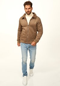 Braune, Reißverschlussjacke mit einem strukturierten, plüschigen Kragen; getragen über einem cremefarbenen Rollkragenpullover, kombiniert mit hellblauen Jeans und weißen Sneakers.