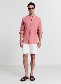 Pink textured button-up shirt, white shorts, and black sandals. Model wears round sunglasses, showcasing a casual summer outfit.