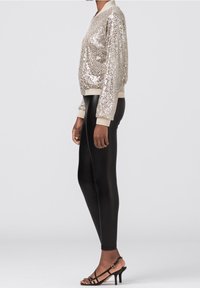 Güldenes Sweatshirt mit Pailletten, beige gerippte Bündchen, kombiniert mit schwarzen Leggings und schwarzen Riemchen-Highheels. Die glatte Textur steht im Kontrast zu den glänzenden Pailletten.