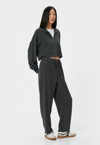 Kort grijs zip-up sweatshirt gecombineerd met losse, gestructureerde grijze broek. Witte shirt eronder en witte sneakers met grijze en zwarte accenten.