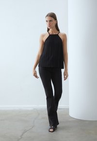 Svart plissert topp med halterneck, parret med flagrende svarte jeans og svarte sandaler med åpen tå. Jevn tekstur; minimalistisk design.