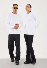 Two people stand wearing white long-sleeve tops; one wears black track pants and sunglasses, the other wears black flared pants and boots.